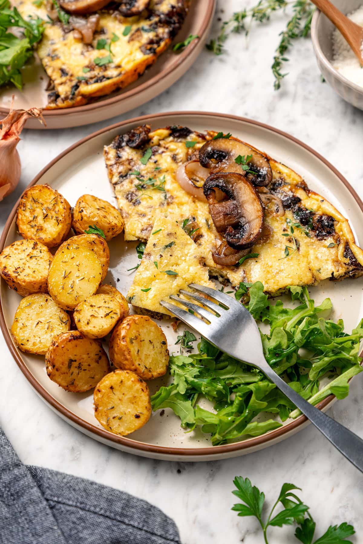 A fork cutting into a slice of mushroom frittata served with crispy potatoes and fresh greens on a plate.