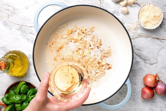 Hand holding wine above skillet with aromatics.