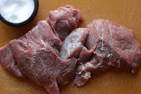 Raw pork shoulder cut into four pieces and seasoned with salt on a countertop.