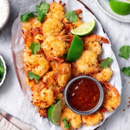 Platter of baked coconut shrimp with cup of sauce and lime wedges.