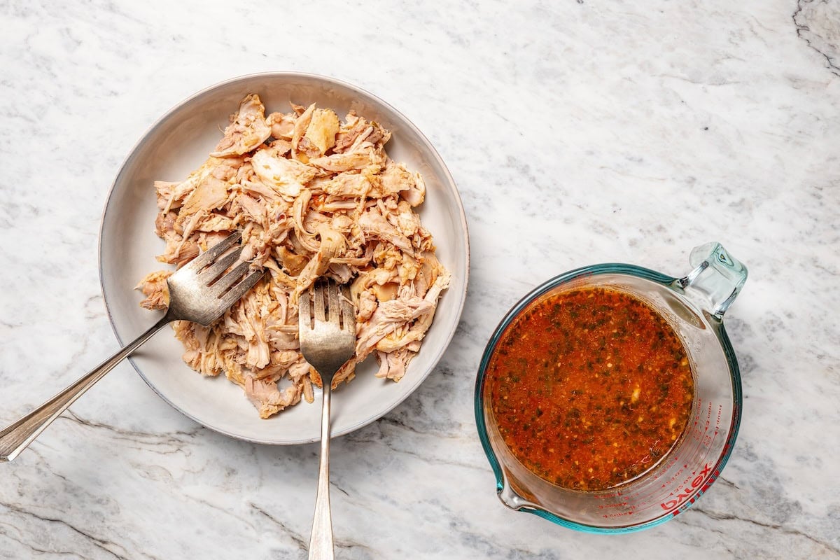 Two forks shredding chicken on a plate next to a cup of cooking liquid.