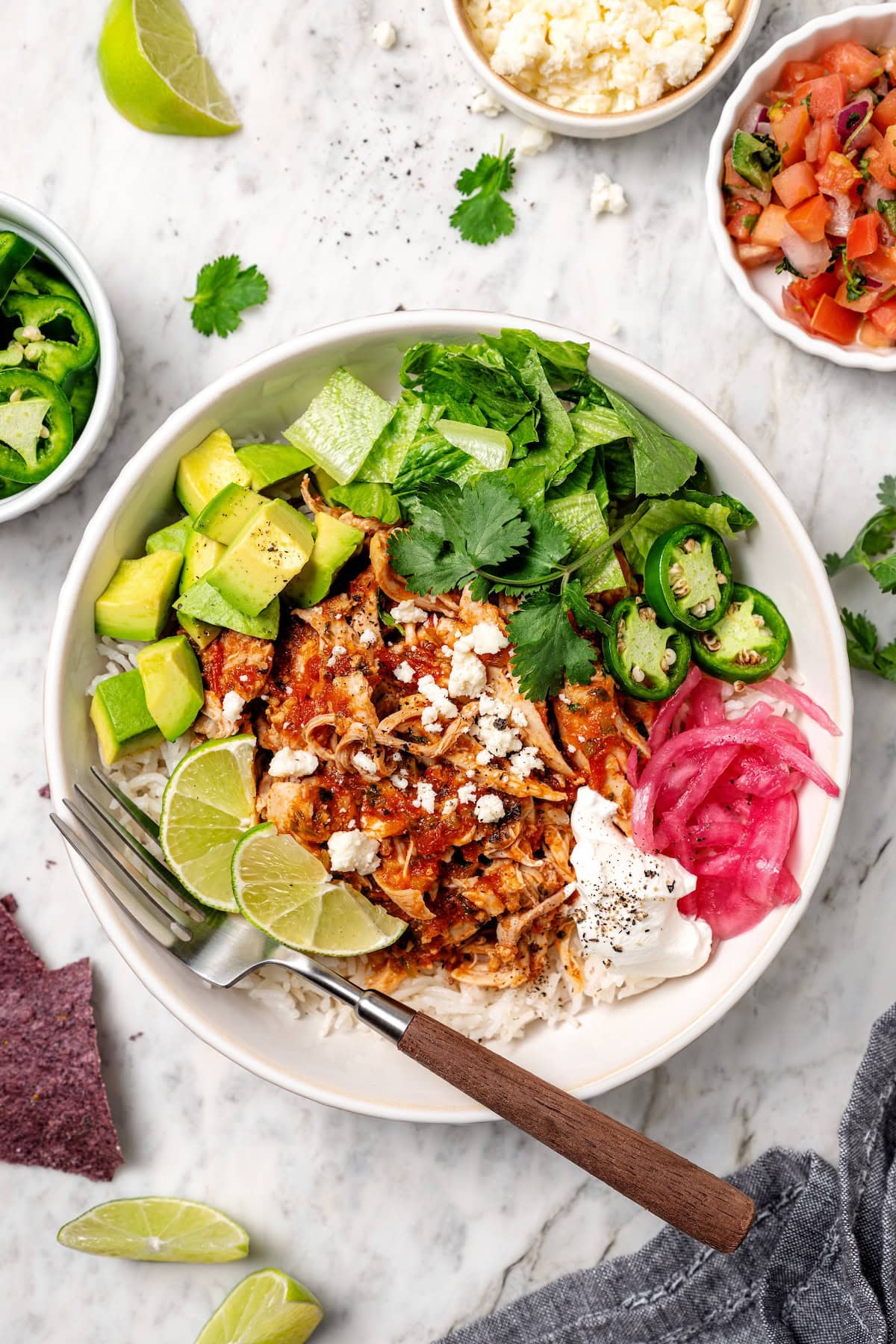 Overhead view of a salsa chicken rice bowl garnished with avocado, pickled onions, jalapeño, lime wedges, and more toppings.
