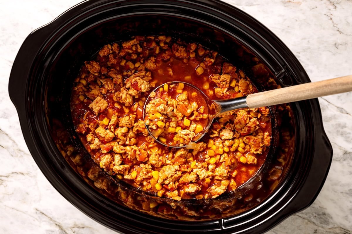 This crock pot turkey chili recipe is easy to make and family-friendly, with ground turkey, corn, bell pepper, tomatoes, and mild spices. Overhead view of a ladle scooping turkey chili inside the crock pot.