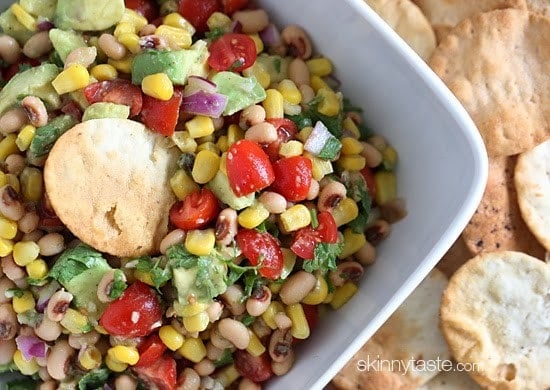 A bowl of black-eyed peas mixed with tomatoes, corn, avocado, and fresh cilantro with a cracker dipping into the bowl