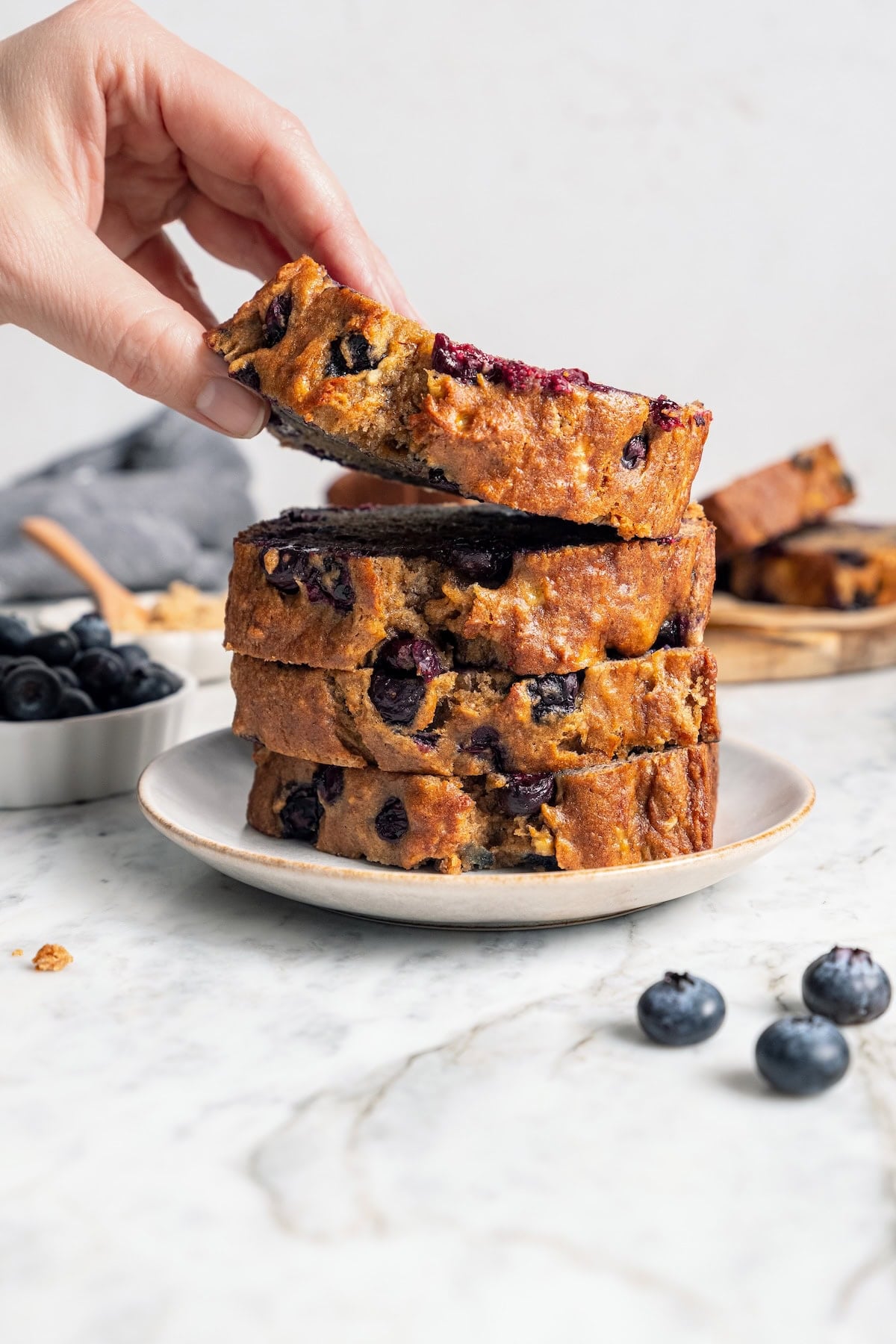 Lifting slice of blueberry banana bread from plate with stack of slices.
