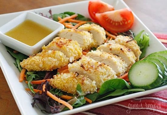 A plate of mixed greens topped with sliced coconut-crusted chicken, with sliced cucumbers, sliced tomatoes and honey mustard dressing on the side.