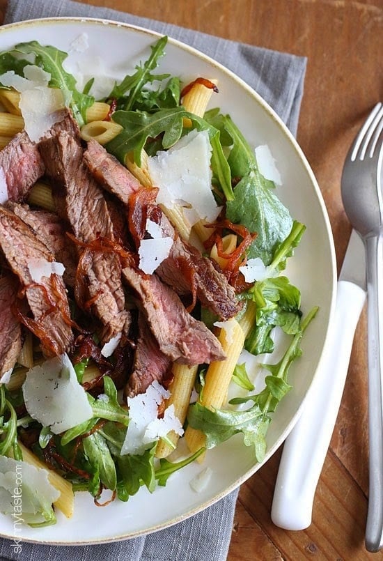 A plate of arugula and penne pasta topped with sliced steak, carmelized onions, and shaved Parmesan cheese.