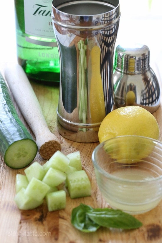 A cocktail shaker, a muddler, a whole cucumber with diced pieces, a whole lemon and a bowl of lemon juice, basil leaves, and a bottle of gin in the background