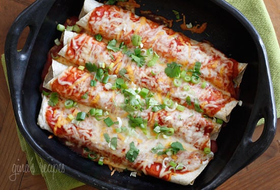 A casserole dish with rolled, filled tortillas covered in enchilada sauce, melted cheese, and sliced scallions.