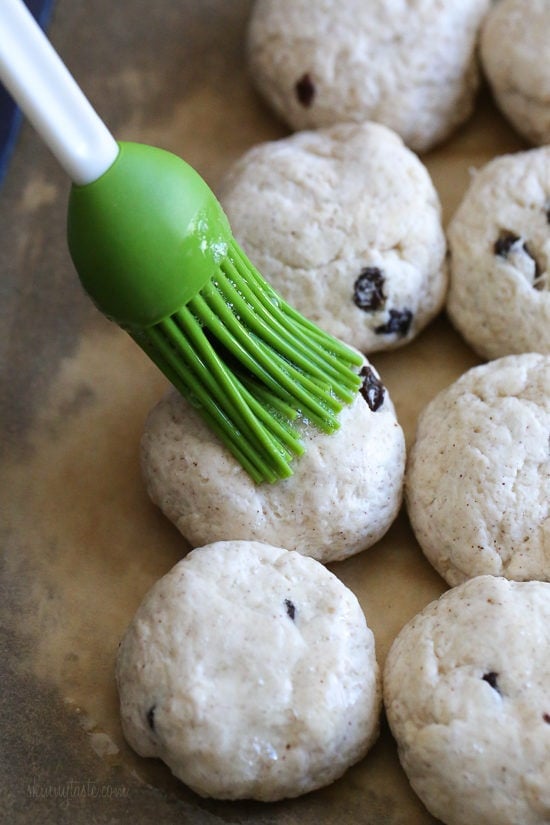 Make soft homemade hot cross buns without yeast, filled with spice, raisins, and finished with icing. Easy to bake in the oven or air fryer! A rubber basting brush brushing egg wash over balls of hot cross bun dough.