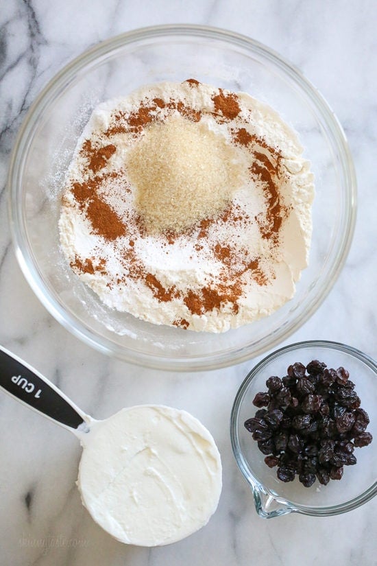Make soft homemade hot cross buns without yeast, filled with spice, raisins, and finished with icing. Easy to bake in the oven or air fryer! A bowl of dry ingredients next to a measuring cup of Greek yogurt an a bowl of raisins.