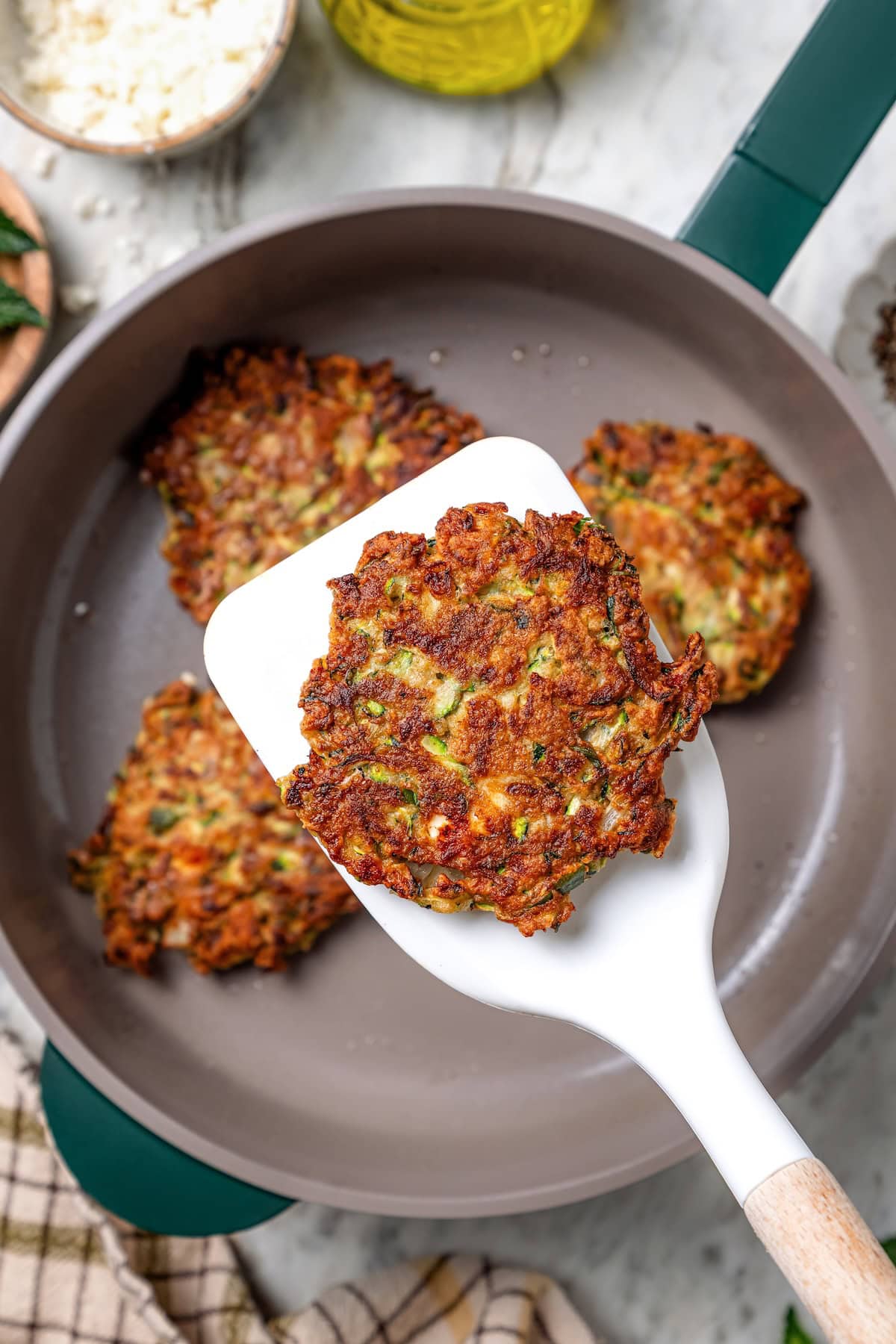 These easy zucchini fritters are crunchy, cheesy, and full of Mediterranean flavor! Serve as a summer side dish, appetizer, or light meal. Spatula for placing zucchini fries on top of a frying pan.