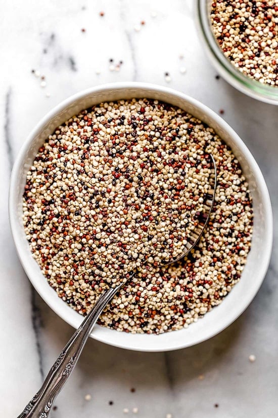 Overhead view of uncooked tricolor quinoa in a white bowl with a spoon for serving.