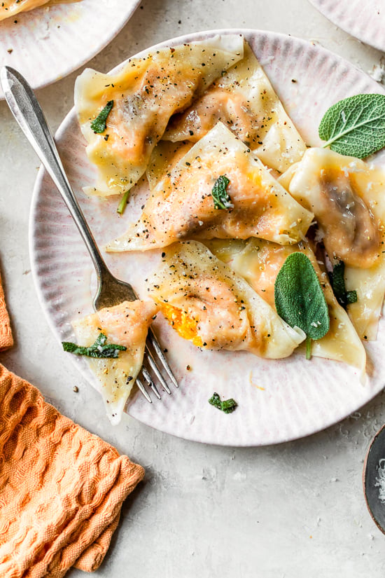 This easy butternut squash ravioli is made with wonton wrappers and tossed with sage butter for a delicious, restaurant-style pasta dinner. A fork cutting into butternut squash ravioli with sage butter on a white plate.