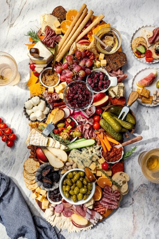 Plates surrounding large charcuterie and cheese board with fruit, pickles, olives, and spreads.
