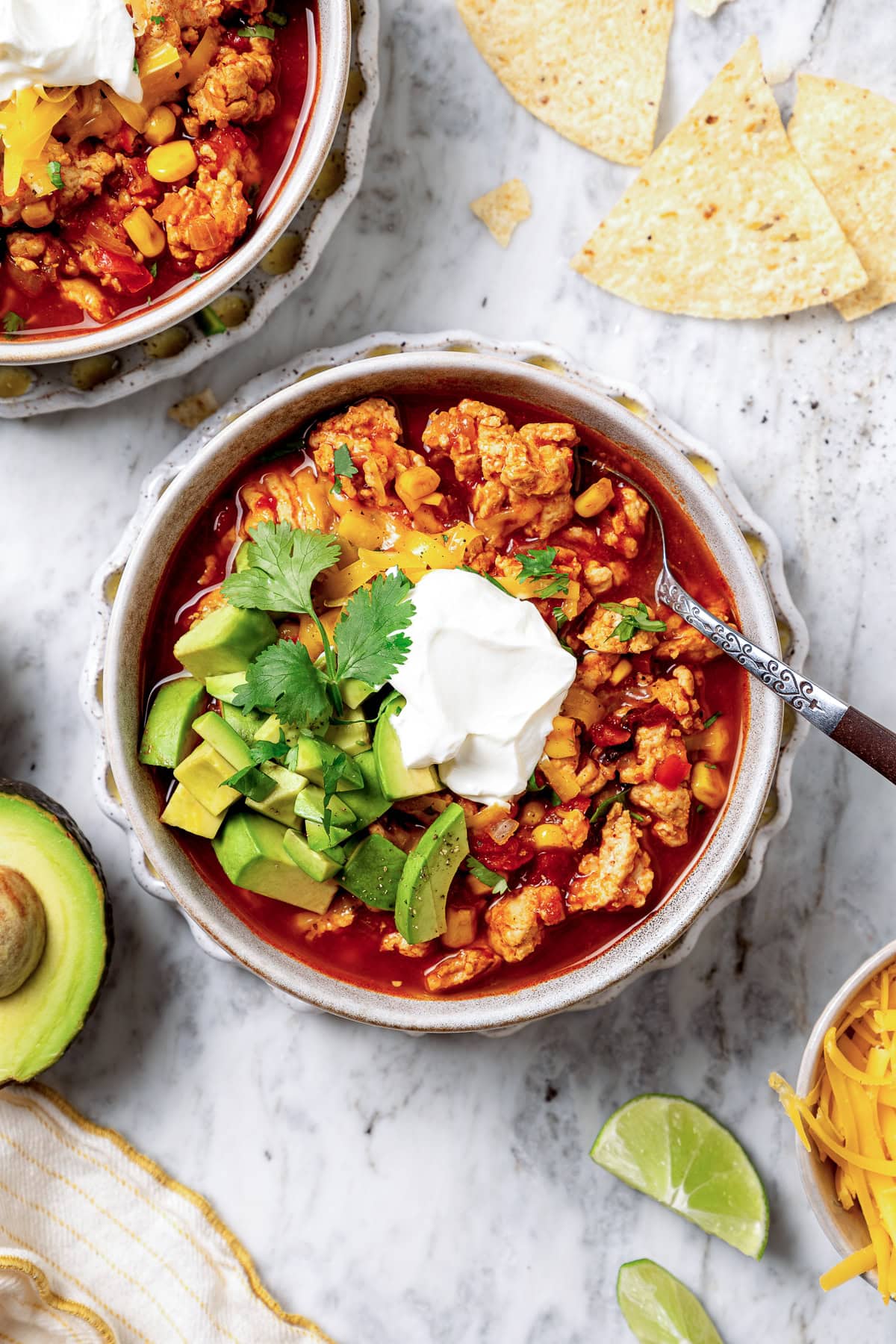 This crock pot turkey chili recipe is easy to make and family-friendly, with ground turkey, corn, bell pepper, tomatoes, and mild spices. A bowl of turkey chili garnished with sour cream, diced avocado, tortilla chips, and cilantro.