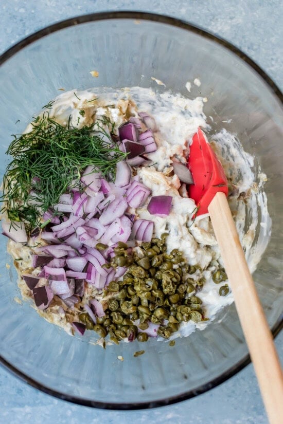 Mixing sardine salad with chopped onions, dill, capers, and mayonnaise in a glass bowl.